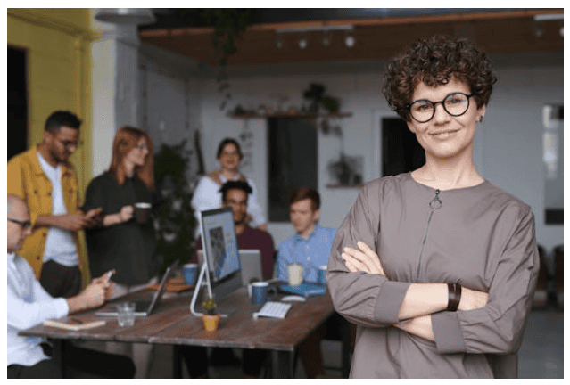 A woman wearing glasses standing confidently with a group of engaged people at the back